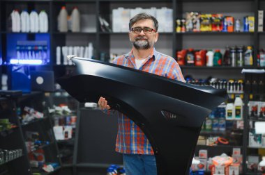 Kendine güvenen bir satıcı, çeşitli araba bakım ürünleriyle çevrili bir oto yedek parça mağazasında yeni bir araba tamponu sunuyor.