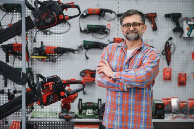 Elektrikli araç gereç dükkanında çapraz pozlar veren kendine güvenen bir satıcı, satılık çok çeşitli ekipmanlar sergiliyor.