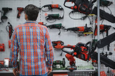Bir hırdavatçıdaki duvarda sergilenen elektrikli testereyi inceleyen müşteri, satın almak için çeşitli seçenekleri göz önünde bulundurarak,