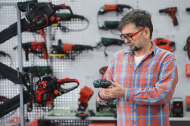 Nalbur dükkanında kablosuz matkabı seçen usta duvarda elektrikli aletler asılı, donanım ve inşaat aletleri için alışveriş yapıyor.