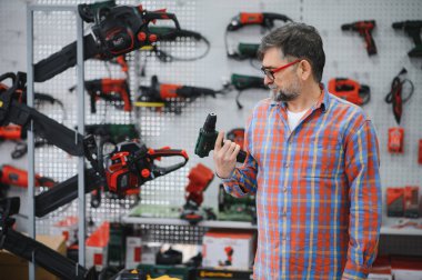 Hırdavatçıda elektrikli matkabı seçen sakallı, olgun bir müşteri. Duvarda çeşitli elektrikli aletler var.