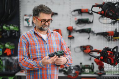 Bir hırdavatçıda kablosuz matkabı inceleyen olgun bir müşteri, duvarda sergilenen elektrikli aletler