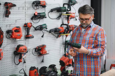Hırdavatçıdaki müşteriye kablosuz matkap gösteren sakallı satıcı, duvarda sergilenen elektrikli aletler