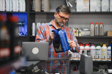 Oto yedek parça mağazası satıcısı motor yağı şişesini barkod tarayıcı ile tarıyor, stoklanmış dükkanda dizüstü bilgisayarla envanteri yönetiyor.