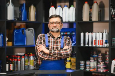 Otomobil yedek parça mağazasında çapraz kollarla poz veren kendinden emin olgun bir satıcı. Etrafı çeşitli araba bakım ürünleriyle dolu raflarla çevrili.