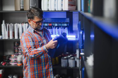 Bir oto yedek parça mağazasında mavi motor yağı taşıyan satıcı etiketi kontrol ediyor ve çeşitli araba bakım ürünleriyle çevrili.