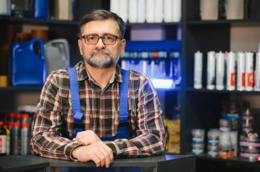 Gözlüklü ve sakallı kıdemli satıcı oto yedek parça dükkanında müşterileri bekliyor.