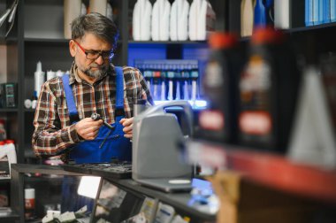 Bir oto yedek parça mağazasında elinde bir alet tutan mavi tulumlu kıdemli satıcı bir ürünü kontrol ediyor.