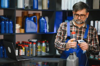 Kıdemli satıcı çeşitli ürünlerle dolu bir oto yedek parça mağazasında çalışırken barkod tarayıcı ile motor yağı şişesini tarıyor.