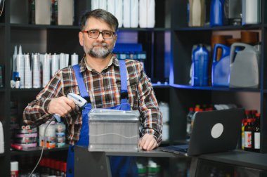 Satış elemanı, otomobil parçaları deposunda barkod tarayıcı ile araba aküsünü tarıyor. Etrafı motor yağı ve diğer ürünlerle çevrili.