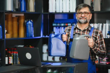 Satıcı, bir oto yedek parça mağazasında farklı motor yağı şişeleri gösteriyor. Rafları ürünlerle dolu ve bilgisayarı tezgahta.