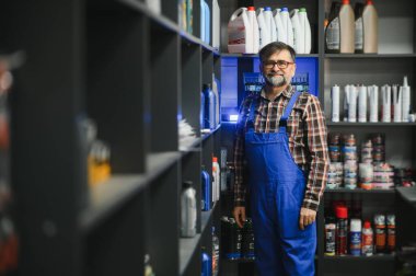 Bir oto yedek parça mağazasında motor yağı, soğutucu ve çeşitli araba bakım ürünleriyle dolu rafların arasında duran kendine güvenen bir satıcı.