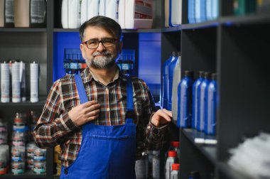Kıdemli satıcı mavi tulumlar giyip rafta motor yağı şişelerini göstererek bir oto yedek parça mağazasında çalışıyor.