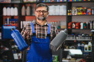 Tamirci, otomotiv malzemeleri raflarıyla çevrili bir oto yedek parça mağazasında çalışırken iki kutu motor yağı tutuyor.