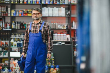 Araba parçaları ve aletler konusunda uzmanlaşmış bir dükkanda mavi tulum giyen bir satıcı elinde motor yağı kutusu tutuyor.