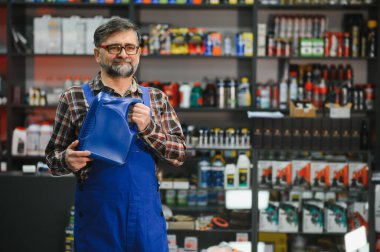 Satıcının elinde bir kutu motor yağı var. Etrafı ürünlerle dolu bir oto yedek parça mağazasında çalışıyor.