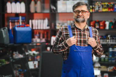 Kendine güvenen mavi tulumlu bir satıcının portresi araba bakım ürünleriyle dolu raflarla çevrili bir oto yedek parça mağazasında duruyor.