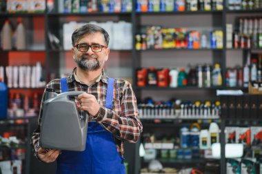 Kıdemli satıcı elinde motor yağı dolu bir teneke kutu tutuyor. Etrafı ürünlerle dolu bir oto yedek parça mağazasında çalışıyor.