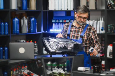 Araba yedek parça mağazası satıcısı, barkod tarayıcısı ile yeni bir far taraması yapıyor. Etrafı araba bakım ürünleriyle dolu.