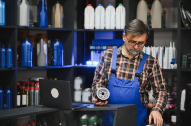 Yeni bir araba yağı filtresi tutan mavi tulumlu kıdemli satıcı ve oto yedek parça mağazasında dizüstü bilgisayarı kontrol ediyor.