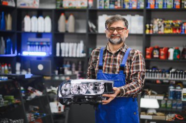 Bir oto yedek parça mağazasında yeni bir far tutan kıdemli satıcı, arka planda bir sürü ürün var.