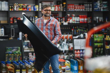 Satıcının elinde araba tamponu var. Etrafı ürünlerle dolu bir oto yedek parça mağazasında müşteriye yardım ediyor.