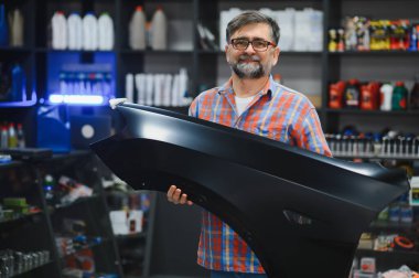 Oto yedek parça dükkanında siyah bir araba çamurluğu tutan kıdemli satıcı müşterilere yardım ve uzmanlık teklif ediyor.