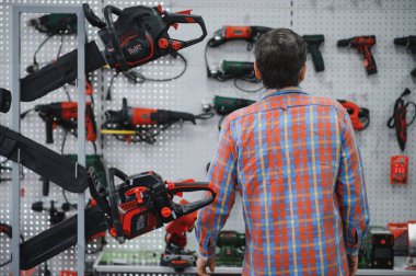 Müşteri hırdavatçıdan elektrikli testereyi seçiyor, duvarda gösterilen elektrikli aletlerin farklı modellerini değerlendiriyor