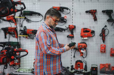 Sakallı, olgun bir adam, çeşitli elektrikli aletler ve ekipmanlarla çevrili, hareketli bir hırdavatçıdan kablosuz bir tornavida seçiyor.