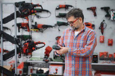 Kıdemli adam bir hırdavatçıda çeşitli elektrikli aletleri karıştırıyor, dikkatle inceliyor ve sergilenen matkapları ve diğer ekipmanları inceliyor