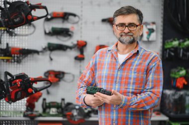 Kıdemli satıcı, çeşitli elektrikli aletler ve ekipmanlarla çevrili bir hırdavatçıda kablosuz matkap sunuyor.