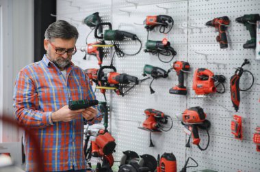 Sakallı olgun müşteri bir hırdavatçıda elektrikli bir araç seçiyor, kutunun üzerindeki özellikleri okuyor.