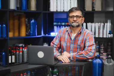 Kendine güvenen olgun bir satıcı, otomobil yedek parça mağazasında dizüstü bilgisayarla duruyor. Etrafı çeşitli araba ürünleriyle çevrili raflarla çevrili.