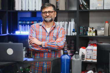 Araba bakım ürünleriyle dolu raflarla çevrili bir oto yedek parça mağazasında çapraz kollu duran bir satıcının portresi.