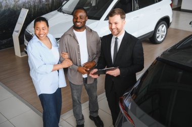 Gülümseyen araba satıcısı, modern bir galeride neşeli bir Afro-Amerikan çifte yeni bir araç sunuyor ve satın alma seçeneklerini tartışıyor.