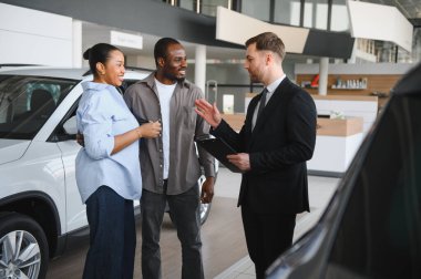 Araba satıcısı elinde dosya ve araba özelliklerini bir araba galerisinde Afro-Amerikan bir çifte anlatıyor.