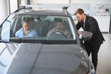 Araba satıcısı, yeni bir arabanın özelliklerini bir araba galerisinde Afro-Amerikan bir çifte sunuyor.