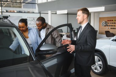 Araba satıcısı, modern bir galeride Afro-Amerikan bir çifte yeni bir arabanın özelliklerini sunuyor.