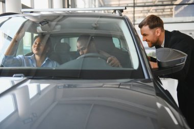 Araba satıcısı, bir galeride sürücü ve yolcu koltuğunda oturan bir çifte yeni bir arabanın özelliklerini gösteriyor.