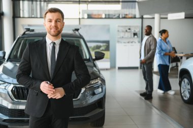 Profesyonel araba satıcısı anahtarlarını tutuyor ve Afrikalı Amerikalı bir çifte modern bir galeride yeni bir araç seçmeleri için yardım ediyor.