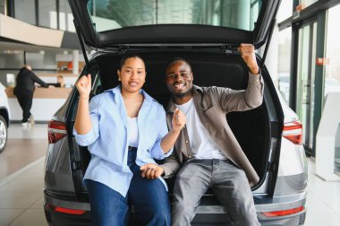 Bir araba bagajında oturan Afrikalı Amerikalı çift yakınlarda yardımsever bir satıcıyla bir galeride yeni araba alımını kutluyorlar.