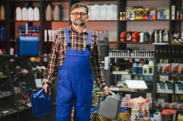 İki motor yağı şişesi tutan mavi tulumlu bir satıcı, bir oto yedek parça mağazasında duruyor, çeşitli ürünlerle dolu raflarla çevrili.