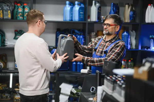 Tamirci araba tamirhanesinde müşteriye araba yağı veriyor, arka planda motor yağlarıyla dolu raflar var.