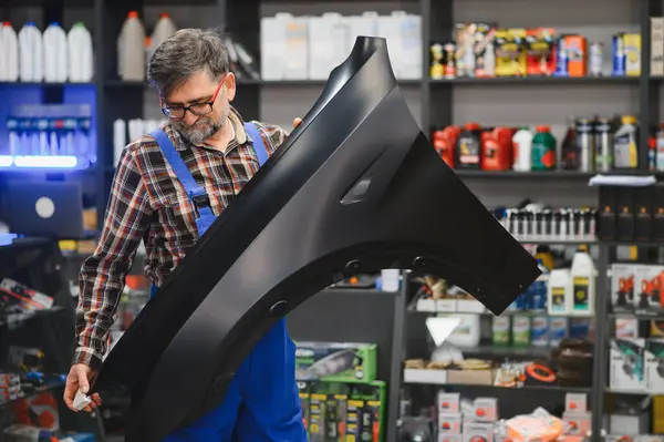 Tamirci, otomobil parçaları satan bir dükkanda içi ürünlerle dolu siyah bir araba tamponu tutuyor.