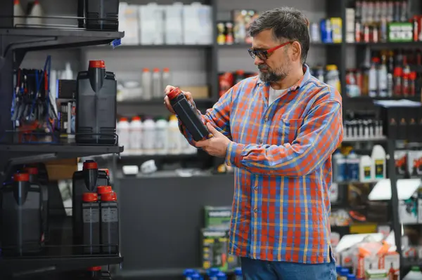 Tamirci oto yedek parça dükkanından motor yağı seçiyor, siyah şişedeki etiketi dikkatle okuyor ve seçeneklerini değerlendiriyor.