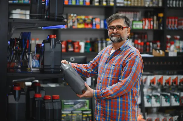 Araba yağı şişesi tutan sakallı, olgun bir satıcı, oto yedek parça mağazasında çalışıyor, müşterilere yardım ediyor.