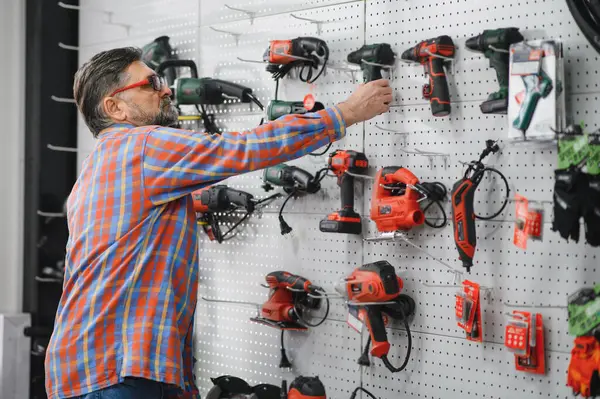 Üst düzey bir adam kablosuz matkap seçerken duvarda sergilenen çeşitli elektrikli aletleri inceliyor.