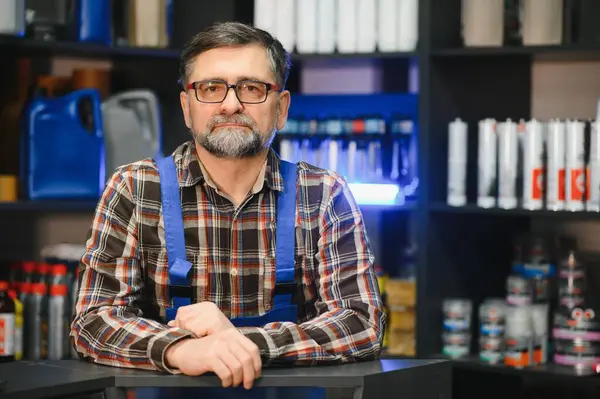 Gözlüklü ve sakallı kıdemli satıcı oto yedek parça dükkanında müşterileri bekliyor.