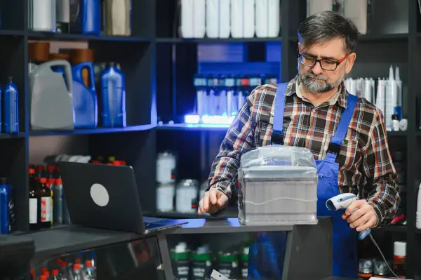 Oto yedek parça mağazası satıcısı araba aküsünü barkod tarayıcı ile tarıyor, dizüstü bilgisayarla envanteri yönetiyor