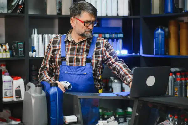 Bir oto yedek parça mağazasında çalışan kıdemli bir satıcı, ürün uygunluğunu kontrol etmek için bir laptop ve barkod tarayıcı kullanıyor.